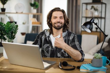 Portrait of attractive male IT specialist typing on keyboard of modern laptop. Young caucasian man sitting at workplace with portable computer and document showing thumb up
