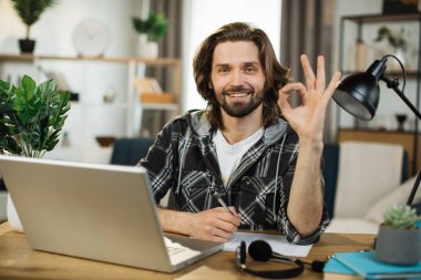 Portrait of attractive male IT specialist typing on keyboard of modern laptop. Young caucasian man sitting at workplace with portable computer and document showing sign ok