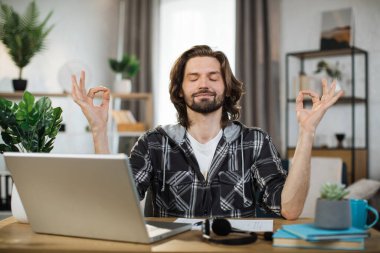 Handsome man freelancer sitting at table with closed eyes and relieving stress by meditation at workplace. Concept of relaxation and harmony, no stress free relief at work.