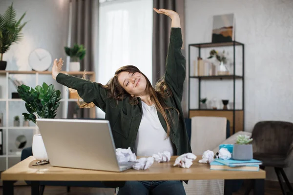 Gorgeous office woman relaxing by stretching her body while sitting in front her computer laptop at the wooden working desk over comfortable living room as background.