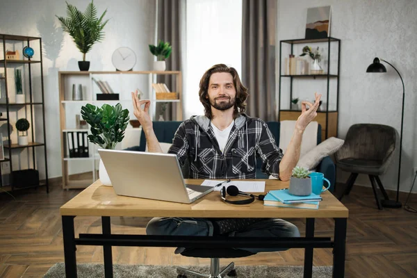 Relaxed caucasian man in casual wear sitting on workplace with modern laptop and meditating with opened eyes. Office worker relief street at work with accomplished fingers.