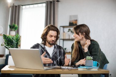 Beautiful caucasian woman standing near bearded man that sitting at table and using laptop and showing information on papers. Two office workers in casual outfit. Work with modern technology.