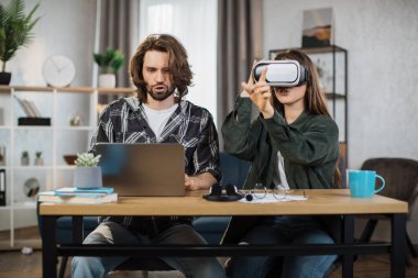 Happy young friends sitting at the table and using modern gadgets for work or study. Pretty girl wearing VR glasses and enjoying artificial reality, while focused man is typing on laptop.