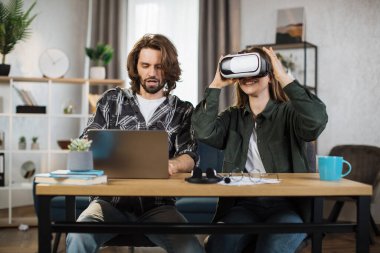 Happy young friends sitting at the table and using modern gadgets for work or study. Pretty girl wearing VR glasses and enjoying artificial reality, while focused man is typing on laptop.