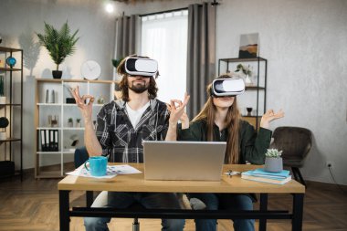Attractive couple sitting together at the table in yoga pose, using VR headsets with modern laptop for experience in virtual reality. Happy family spending leisure time at home and playing games