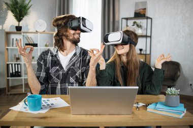 Young couple in casual wear using VR headset and laptop for playing games, sitting in yoga pose during free time at home. Concept of family and entertainment.