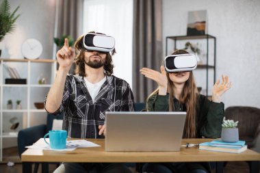Genç çift, sanal gerçeklik deneyimi için modern dizüstü bilgisayarlı VR kulaklıkları kullanarak masada birlikte oturuyorlar. Mutlu aile evde vakit geçirip oyun oynuyor..