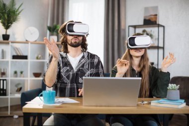 Genç çift, sanal gerçeklik deneyimi için modern dizüstü bilgisayarlı VR kulaklıkları kullanarak masada birlikte oturuyorlar. Mutlu aile evde vakit geçirip oyun oynuyor..