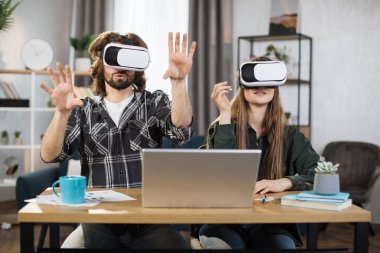 Genç çift, sanal gerçeklik deneyimi için modern dizüstü bilgisayarlı VR kulaklıkları kullanarak masada birlikte oturuyorlar. Mutlu aile evde vakit geçirip oyun oynuyor..