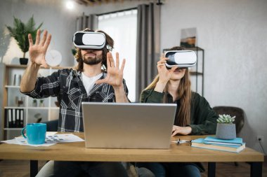 Excited young people with surprised expression, playing games at home, using VR glasses and laptops. Happy family enjoying artificial reality entertaining themselves with innovations.