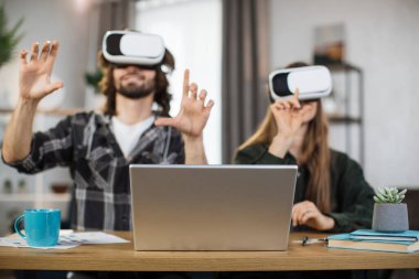 Excited young people with surprised expression, playing games at home, using VR glasses and laptops. Happy family enjoying artificial reality entertaining themselves with innovations.
