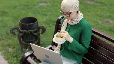 İşteyken sağlıklı salata yiyen çekici Müslüman iş kadını. Şehir parkında ofis binasının yanındaki bankta otururken salatasını yiyen nazik, İslamcı bir kadın..