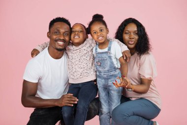 Happy loving family. Close up portrait of african american man, woman and two girls sitting on the floor isolated on pink studio wall. Smiling daughters emracing their mum and dad, posing at camera.