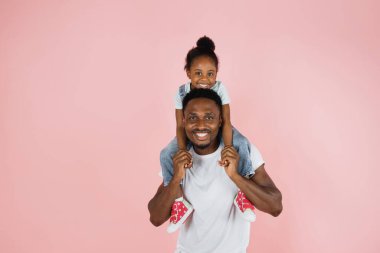 Happy family. Portrait of cheerful african american man riding excited daughter on his shoulders, looking and posing at camera, having fun, holding hands isolated over pink studio background.