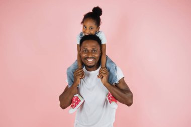 Happy family. Portrait of cheerful african american man riding excited daughter on his shoulders, looking and posing at camera, having fun, holding hands isolated over pink studio background.