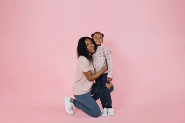 Indoor studio shot of pleasant young African woman mother and her cute adorable little daughter, wearing bright trendy casual clothes, posing together on pink background.