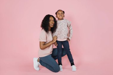 Indoor studio shot of pleasant young African woman mother and her cute adorable little daughter, wearing bright trendy casual clothes, posing together on pink background.