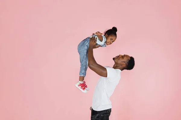Happy fatherhood. Cheerful african dad holding his cute daughter on hands pretending she are flying, girl spreading hands, imitating plane, pink studio background.