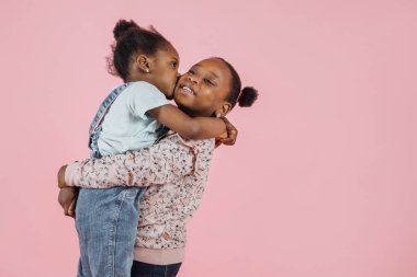 Two cute African girls sisters in casual summer outfits, hugging each other and one girl kissing other in her cheek on pink studio background, copy space.