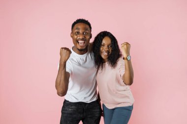 Big win. Happy excited african couple celebrating success with raised fists. Cheerful african american man and woman exclaiming with joy while standing isolated over pink background, copy space.