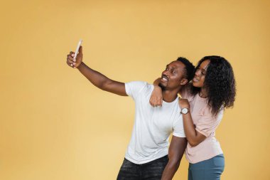 Cheerful african american millennial couple using cell phone for doing picture of themselves on yellow background. Charming women hugging her boyfriend back while he doing selfie, copy space.