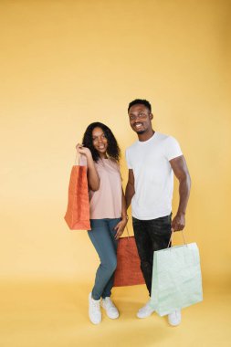 Couple of shopaholics. African american man and woman holding shopping bags, enjoying new goods, yellow studio background, copy space.
