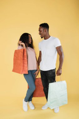 Couple of shopaholics. African american man and woman holding shopping bags, enjoying new goods, yellow studio background, copy space.