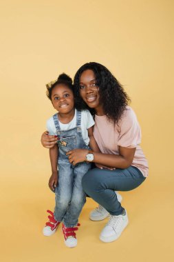 Indoor studio shot of pleasant young African woman mother and her cute adorable little daughter, wearing bright trendy casual clothes, posing together on yellow background.