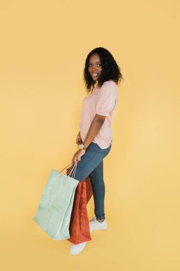 Pleasant young African woman in pink t-shirt, enjoying shopping, posing to camera over isolated yellow background with many colorful shopping bags. Close up, copy space.