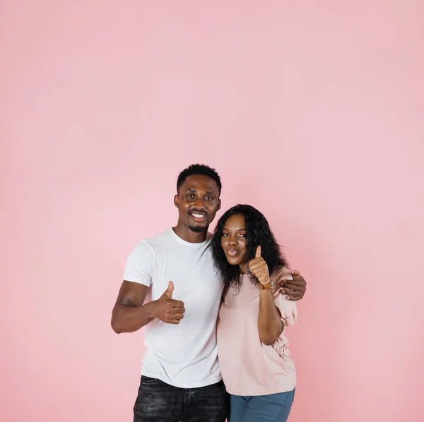 Photo below belt of cheerful people of exotic appearance on isolated pink wall. African female with her arms on waist dressed in pink T-shirt and jeans while guy in white t-shirt showing thumb up