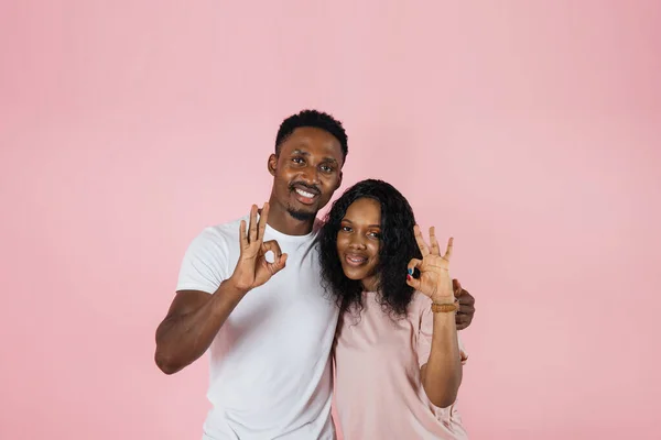 Photo below belt of cheerful people of exotic appearance on isolated nacre pink wall. African female with her arms on waist dressed in pink T-shirt and jeans while guy in white t-shirt showing sign ok