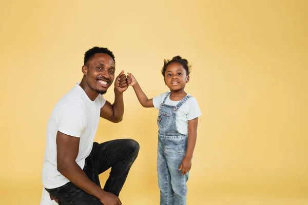 Smiling african american dad and his daughter girl in casual clothes posing isolated on yellow background studio portrait. People emotions lifestyle concept. Mock up copy space. Giving fists bump.