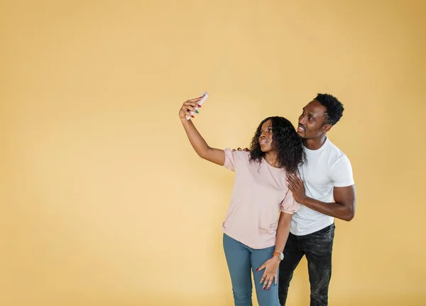 Joyful people using mobile phone to take selfies together in studio. African man and woman taking picture with smartphone and having fun in front of camera. Carefree couple feeling happy.