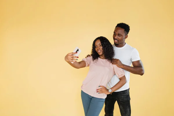 Joyful people using mobile phone to take selfies together in studio. African man and woman taking picture with smartphone and having fun in front of camera. Carefree couple feeling happy.