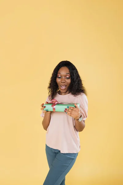 Portrait of excited funny african american woman with afro hair, wearing pink t-shirt, posing with gift box holding in her hand, isolated on yellow background.