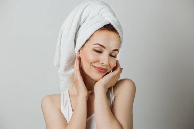 Caucasian young lady in white towel touching gently her cheek, smiling with closed eyes. Beautiful woman with bare shoulders posing over grey background.