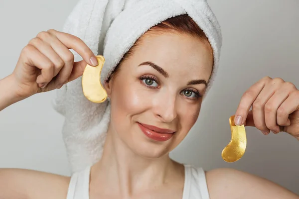 Portrait of pleasant caucasian young woman in towel with healthy skin holding golden eye patches in hands and smiling on camera. Isolated over grey studio background.