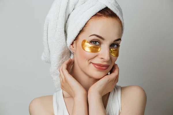 Portrait of caucasian young woman in towel touching with her hands golden hydrogel patches under eyes. Isolated over grey studio background.