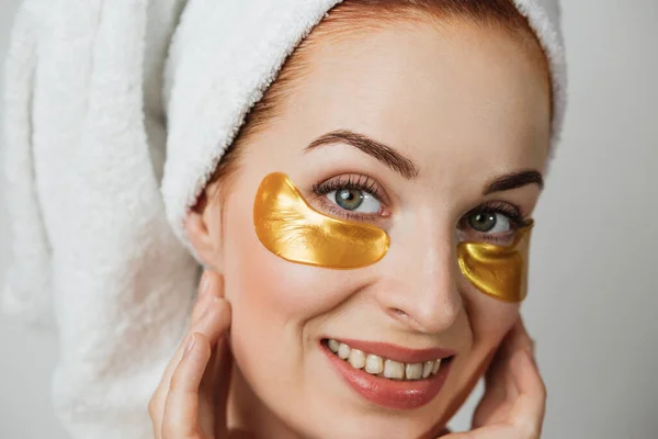 Close up face of adorable young woman in towel after shower, using golden collagen patches for smoothing skin under eyes. Concept beauty treatments.