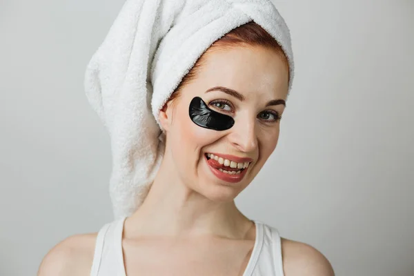 Portrait of pleasant young woman in towel posing in studio with collagen black patch under eye showing her tongue. Charming female lady using cosmetic for beauty care.