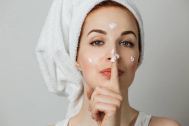 Caucasian young woman using moisturising cream for smoothing skin on face. Adorable female showing sign of silence over grey background.