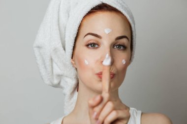 Caucasian young woman using moisturising cream for smoothing skin on face. Adorable female showing sign of silence over grey background.