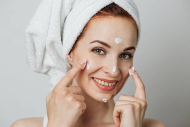 Portrait of caucasian young lady applying face cream over grey background. Adorable woman smoothing skin under eyes with professional cosmetic.