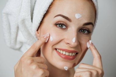 Portrait of caucasian young lady applying face cream over grey background. Adorable woman smoothing skin under eyes with professional cosmetic.