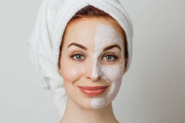 Attractive young woman posing in studio with white mask on half of her face. Caucasian lady in towel using cosmetic for doing anti aging procedures.