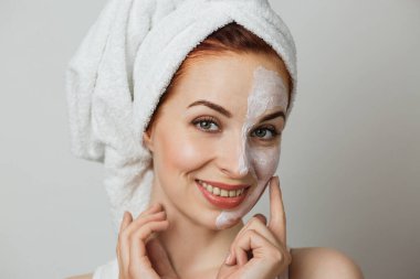 Attractive young woman posing in studio with white mask on half of her face. Caucasian lady in towel using cosmetic for doing anti aging procedures.