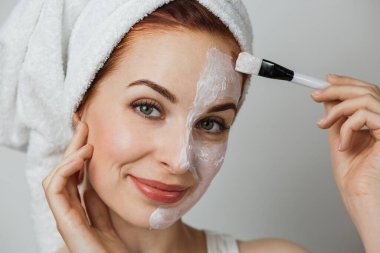 Caucasian young woman in towel using brush for applying white cosmetic cream on half of face over grey studio background. Close up, copy space.