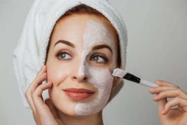 Portrait of caucasian young woman with using brush for applying facial skincare mask posing over grey background. Attractive female with professional cosmetic in studio.