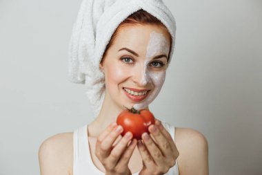 Beautiful young woman with cosmetic mask on half of face and towel holding tomato over grey background. Pretty lady doing skin care procedures in studio.