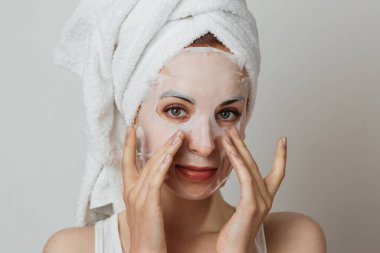 Stunning caucasian lady in towel applying paper mask sheet on face in studio. Young woman enjoying skin care procedures over grey background.